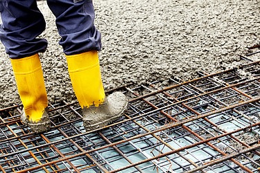 Baustelle mit Betongebäuden, Stahlträgern und Bauarbeitern mit Sicherheitsausrüstung unter teils bewölktem Himmel