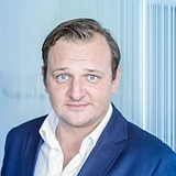 Das Bild zeigt eine Person mit kurzem, leicht gewelltem Haar und blauen Augen. Sie trägt einen blauen Anzug und ein weißes Hemd. Der Hintergrund ist unscharf und besteht aus vertikal verlaufenden Elementen, die auf eine moderne Innenarchitektur hindeuten.