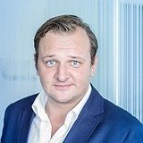 Das Bild zeigt eine Person mit kurzem, leicht gewelltem Haar und blauen Augen. Sie trägt einen blauen Anzug und ein weißes Hemd. Der Hintergrund ist unscharf und besteht aus vertikal verlaufenden Elementen, die auf eine moderne Innenarchitektur hindeuten.