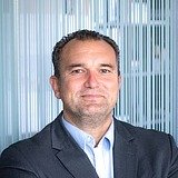 Das Bild zeigt einen Mann mittleren Alters mit kurzem, dunklem Haar. Er trägt ein dunkelgraues Sakko und ein hellblaues Hemd. Der Hintergrund besteht aus vertikalen, halbtransparenten Streifen. Der Mann blickt leicht lächelnd in die Kamera.