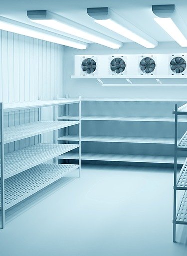 Leerer Kühlraum mit glattem, hygienischem Betonboden und seitlichen Metallregalen; am Ende des Raums befinden sich vier große Kühlventilatoren.