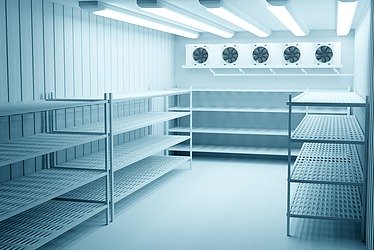 Leerer Kühlraum mit glattem, hygienischem Betonboden und seitlichen Metallregalen; am Ende des Raums befinden sich vier große Kühlventilatoren.