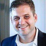 Ein Mann in Business-Kleidung steht vor einer Glastür. Er trägt ein weißes Hemd und ein dunkelblaues Sakko, hat kurzes dunkles Haar und blickt freundlich in die Kamera.