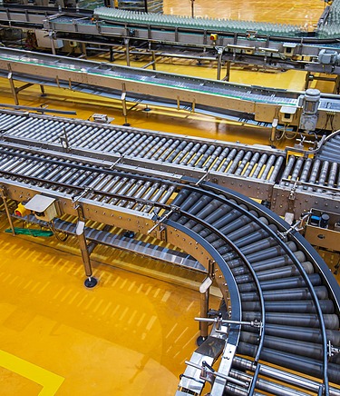 Förderbandsystem in einer Industriehalle mit gelbem Boden und metallischen Maschinenkomponenten.