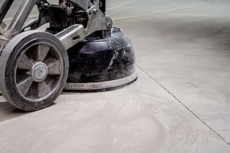 Boden-Schleifmaschine beim Schleifen einer Betonoberfläche. Auf dem Boden sind kreisförmige Schleifspuren und Staubrückstände zu sehen, die vom Arbeitsvorgang stammen.