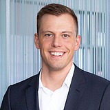 „Das Bild zeigt einen Mann mit kurzem Haar, der einen dunklen Anzug und ein weißes Hemd trägt. Er steht vor einer Wand mit vertikal verlaufenden Streifen und blickt lächelnd in die Kamera.“