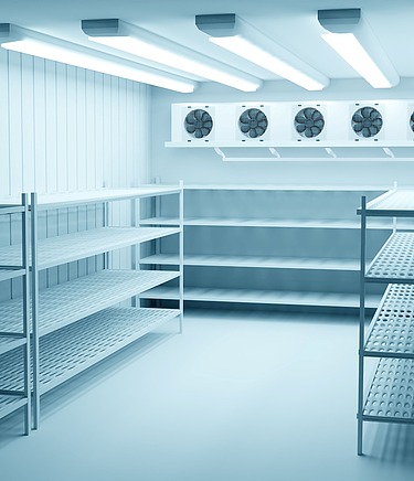 Leerer Kühlraum mit glattem, hygienischem Betonboden und seitlichen Metallregalen; am Ende des Raums befinden sich vier große Kühlventilatoren.