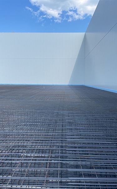 Bewehrte Betonbodenfläche mit dicht verlegter Stahlarmierung in einer modernen Industriehalle vor dem Betonieren, großflächiger Rohbau mit hellen Betonwänden und blauem Himmel