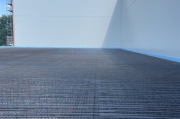Bewehrte Betonbodenfläche mit dicht verlegter Stahlarmierung in einer modernen Industriehalle vor dem Betonieren, großflächiger Rohbau mit hellen Betonwänden und blauem Himmel