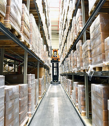 Innenansicht eines Hochregallagers mit palettierten Kartons in deckenhohen Regalen. Ein Gabelstapler steht im Mittelgang auf einem glatten, tragfähigen Betonbode