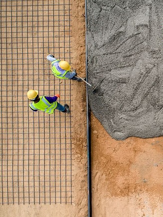 Luftaufnahme von Bauarbeitern beim Verteilen und Glätten von frischem Beton auf einer Baustelle, mit sichtbarer Bewehrung und Sanduntergrund – Betonbodenbau für Fundamentarbeiten.