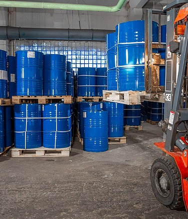 Gabelstapler hebt zwei blaue Fässer auf einer Palette in einer Lagerhalle mit flüssigkeitsdichtem Betonboden für LAU-Anlagen.