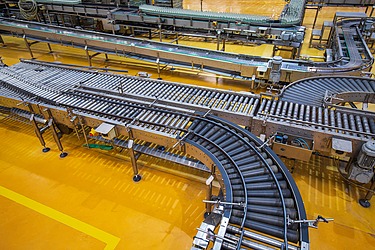 Förderbandsystem in einer Industriehalle mit gelbem Boden und metallischen Maschinenkomponenten.